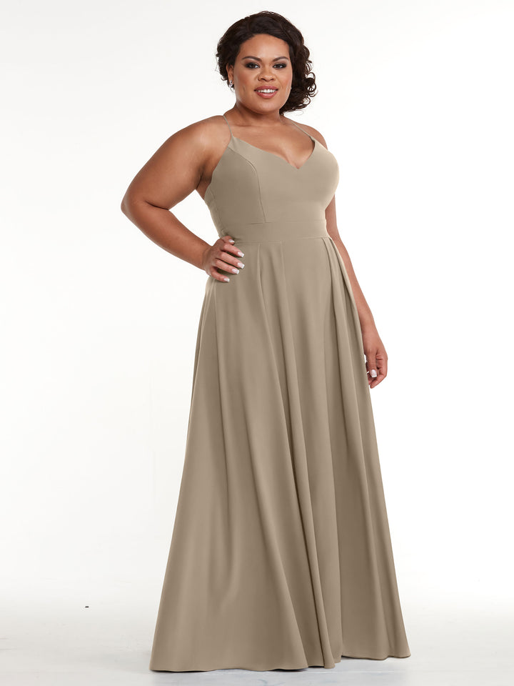 A woman wearing Avery Austin’s 83027, a sleeveless V-neck taupe A-line dress with a fitted bodice and flowing skirt, stands against a white background, smiling confidently.