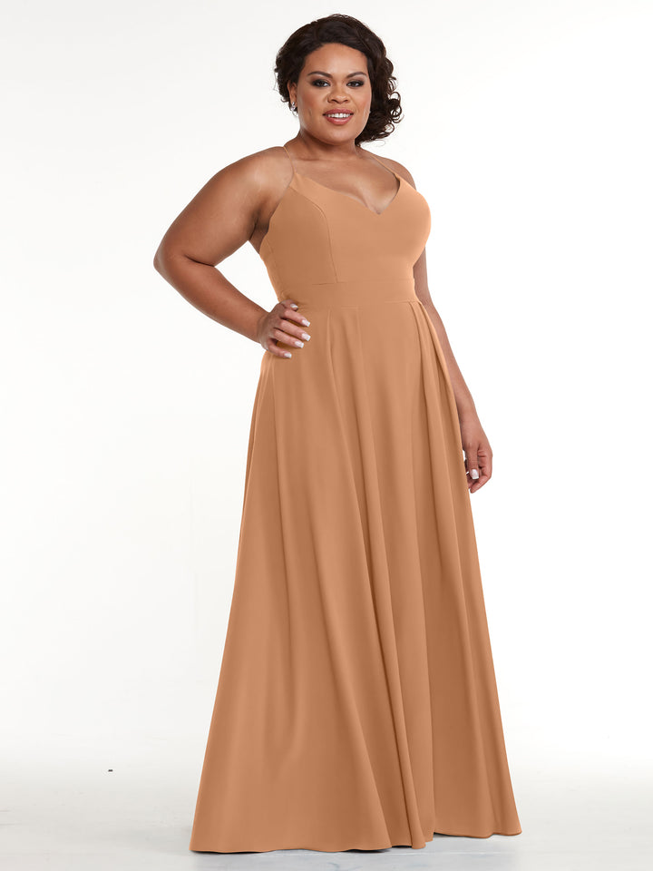 A woman smiles with one hand on her hip, modeling the Avery Austin 83027 floor-length sleeveless camel A-Line dress with a V-Neck, standing against a plain white background.