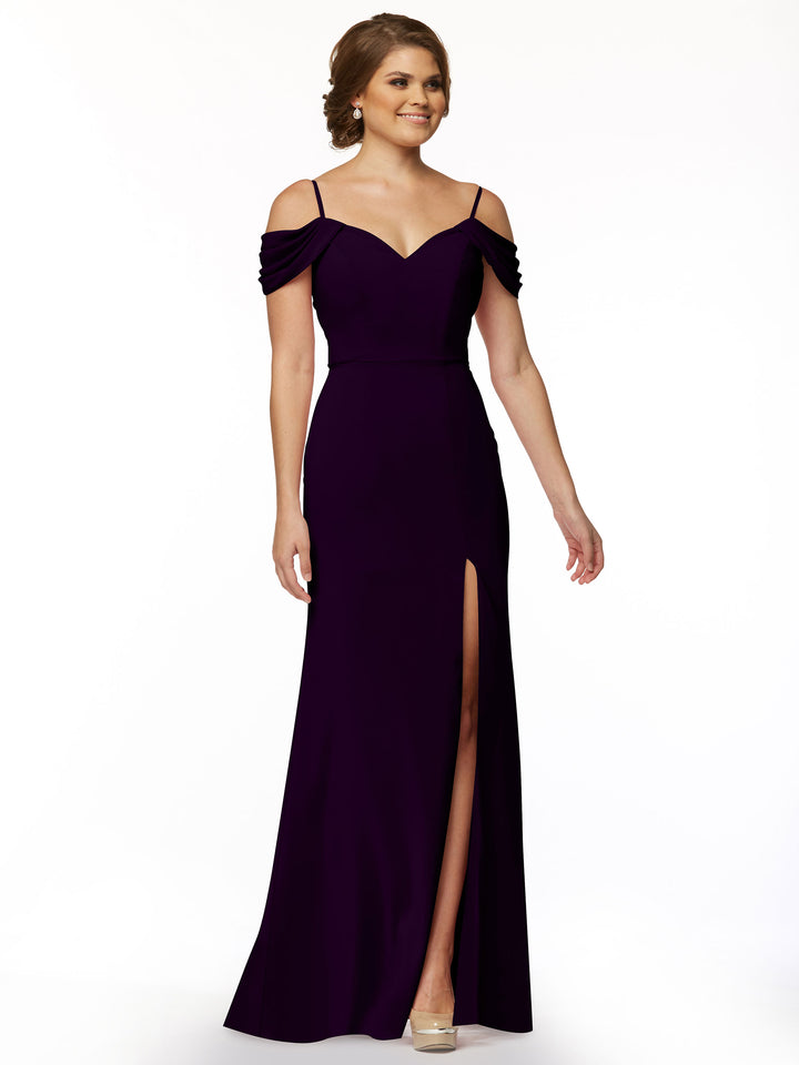 A woman in Avery Austin’s floor-length, fitted off-the-shoulder dress (style 83029) with spaghetti straps and a thigh-high slit poses against a white background. She has short brown hair and beige high heels.