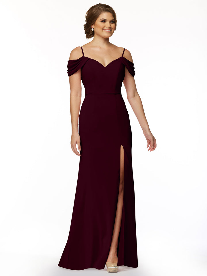 A woman wearing Avery Austin’s 83029, a floor-length, fitted dress with off-the-shoulder spaghetti straps and a high thigh slit, stands smiling against a plain white background.