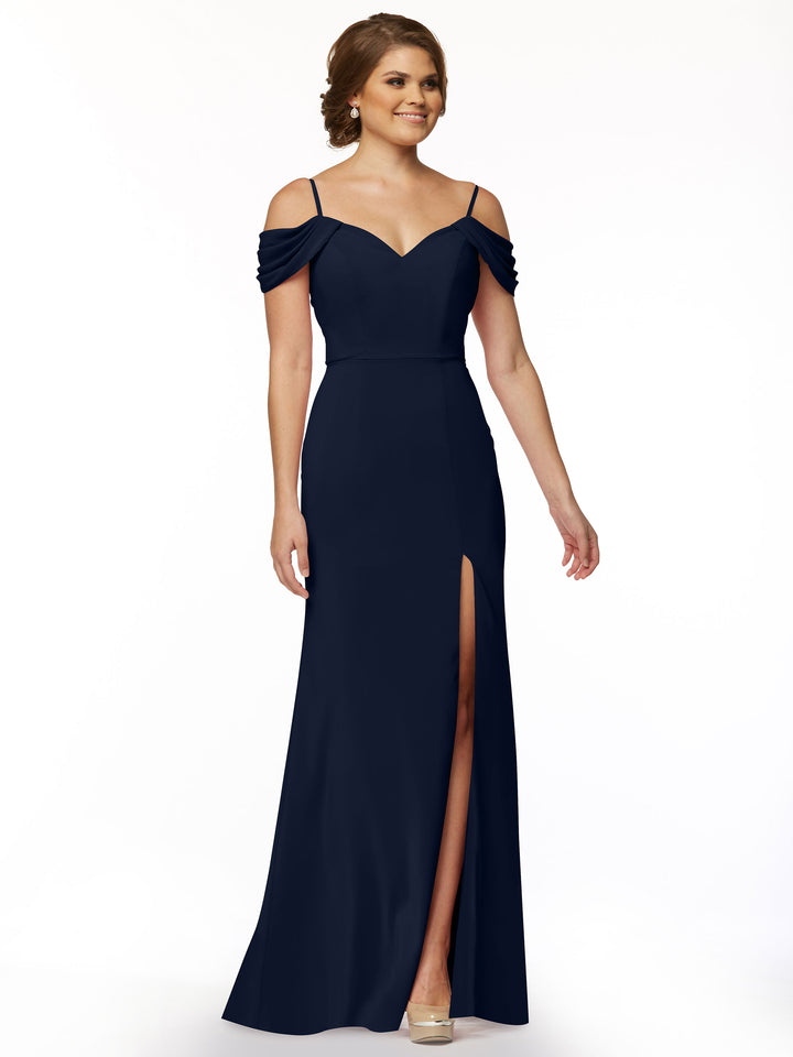 A woman smiles against a white background wearing Avery Austin style 83029, a navy off-the-shoulder dress with spaghetti straps, sweetheart neckline, draped sleeves, fitted bodice, and thigh-high slit.