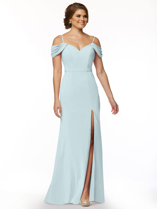 A woman in Avery Austin's sky blue off-the-shoulder dress (style 83029), featuring draped sleeves and a thigh-high slit, stands against a plain white background with her short brown hair styled up.