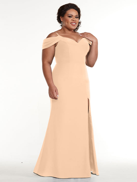 A woman smiles and gently touches her shoulder while modeling the Avery Austin TH-83029 off-the-shoulder peach dress with a front slit, posed against a plain white background.
