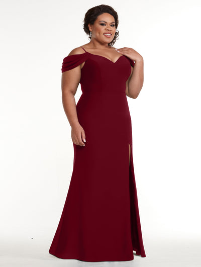 A woman stands and smiles in the Avery Austin 83029, a floor-length fitted off-the-shoulder burgundy dress with a slit, posed against a plain white background.