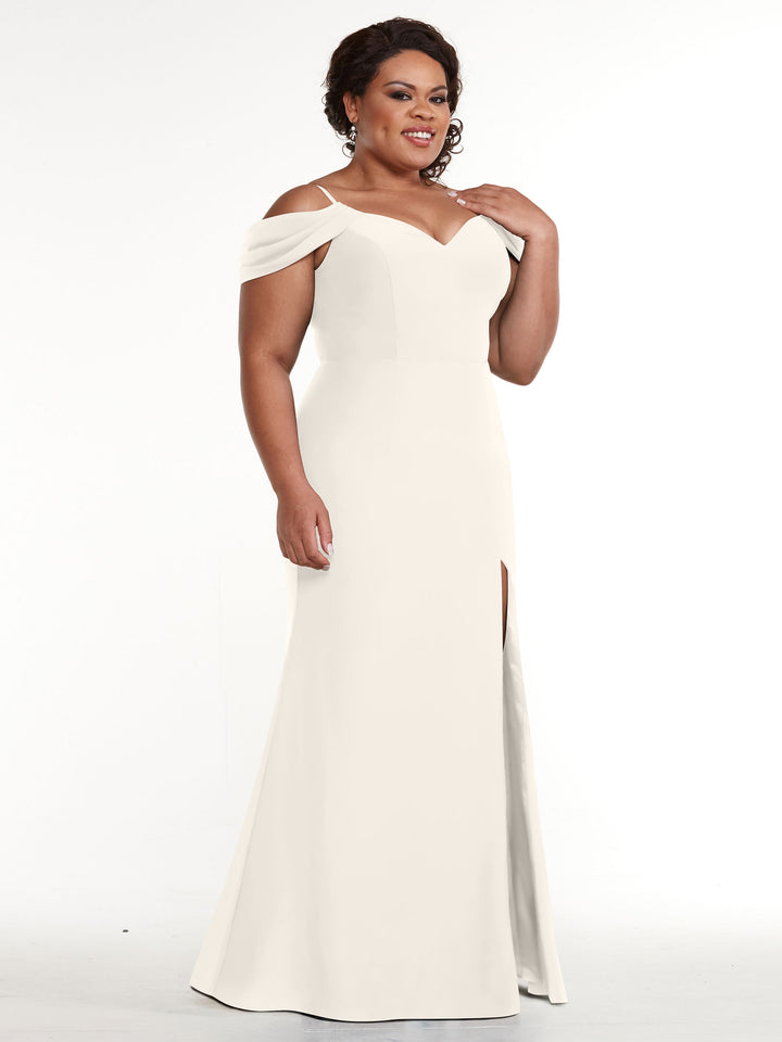 A woman smiles against a plain white background wearing Avery Austin’s 83029: an off-the-shoulder, floor-length white gown with a side slit and fitted silhouette.