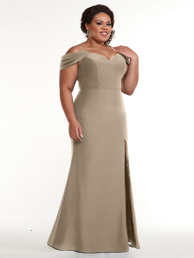 A woman models Avery Austin’s 83029, an elegant light brown off-the-shoulder dress with a thigh-high slit, standing confidently against a plain white background.