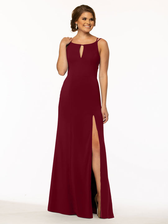 A woman stands smiling against a plain white background, wearing Avery Austin style 83030—a sleeveless, floor-length burgundy sheath dress with a front keyhole cutout and high slit skirt.