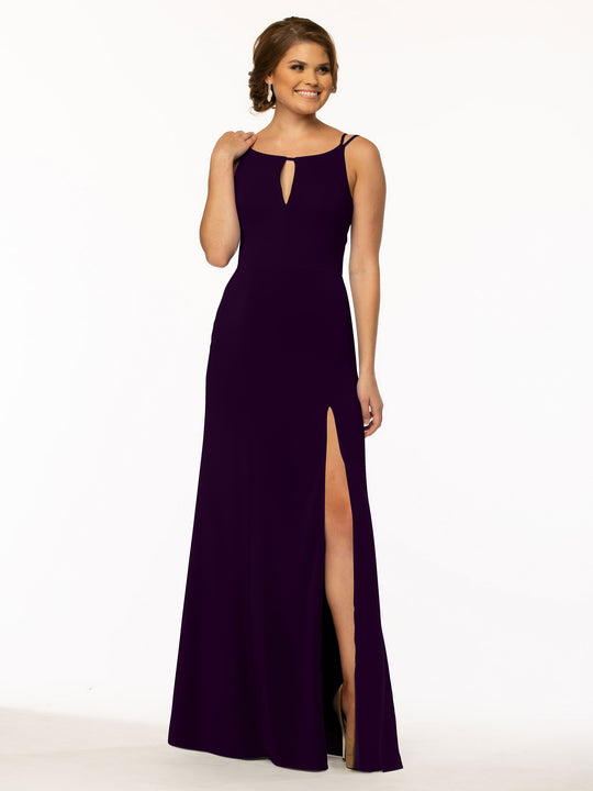 A woman stands and smiles in front of a plain white background wearing the Avery Austin 83030: a sleeveless, floor-length, dark purple sheath dress with a front keyhole cutout and high slit skirt.