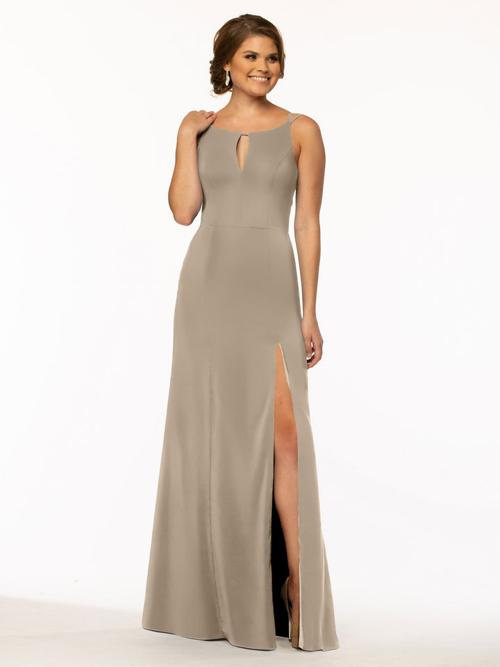 A woman smiles against a plain white background wearing Avery Austin style 83030, a sleeveless taupe sheath dress with a bateau neckline, front keyhole cutout, and high-slit skirt.