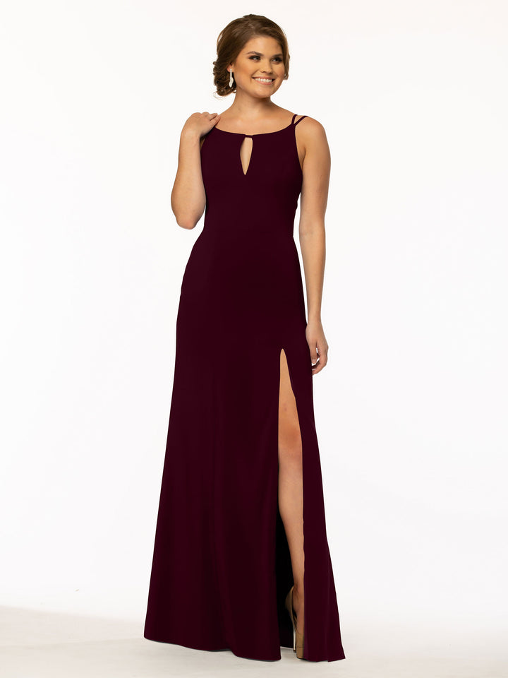 A woman stands and smiles against a plain white background, wearing Avery Austin style 83030—a sleeveless, floor-length dark burgundy sheath dress with a keyhole neckline and high slit.