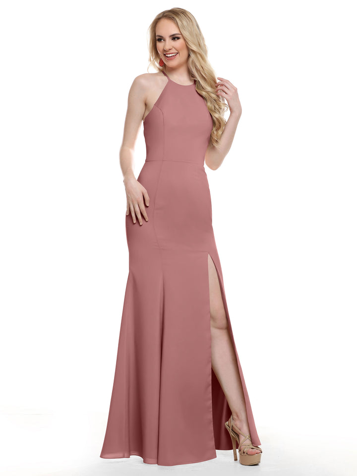 A woman stands smiling in Avery Austin’s 83031, a sleeveless rose halter gown with a high neckline and thigh-high slit, one hand on her wavy blonde hair and the other on her hip, paired with high-heeled shoes.