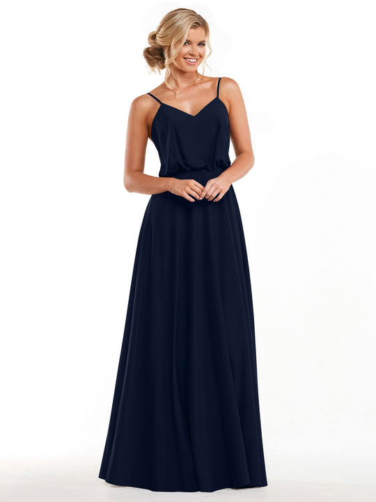 A smiling woman with blonde hair in an updo wears Avery Austin's 83040—a sleeveless, floor-length navy blue A-line dress with thin straps and a V-neckline—standing with hands clasped against a plain white background.