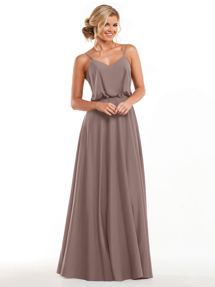 A woman smiles with her hands clasped while modeling Avery Austin’s 83040, a long taupe A-line dress with thin straps and a flowy skirt, against a plain white background.