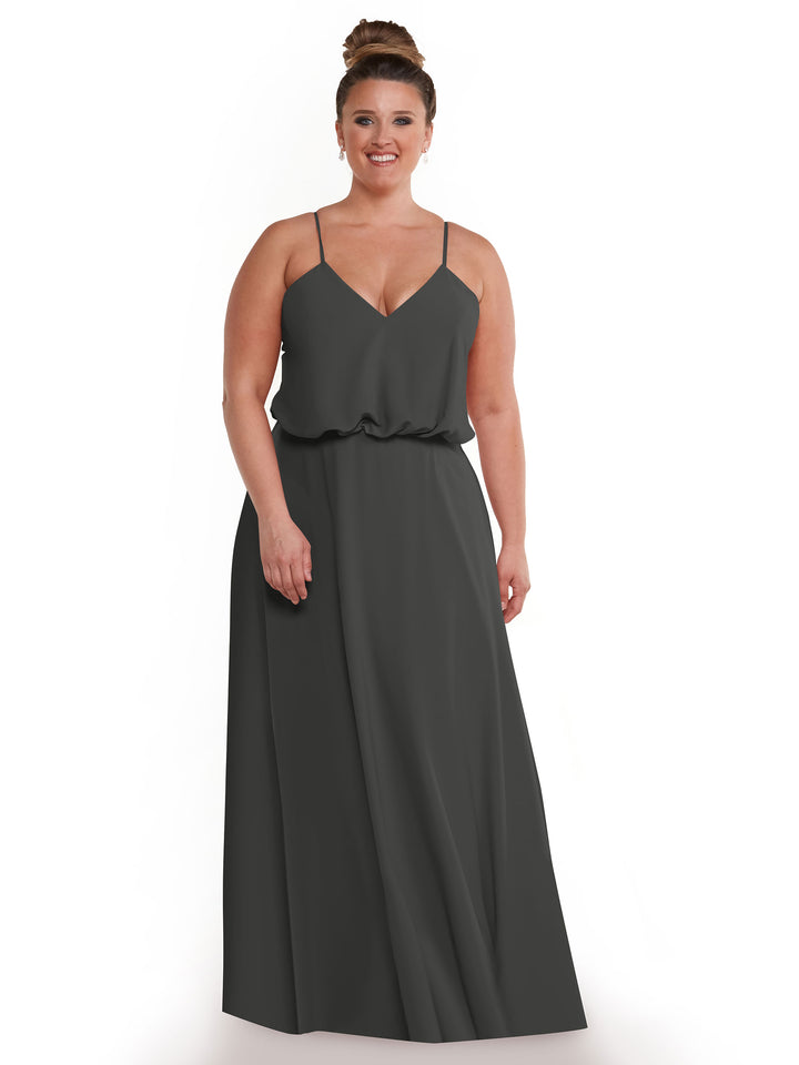 A woman with light skin and brown hair in a bun wears Avery Austin style 83040—a dark gray, sleeveless A-line dress with thin straps, V-neckline, and flowy skirt—standing and smiling against a white background.