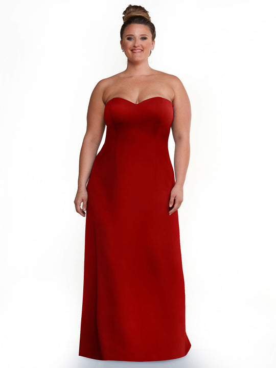 A woman with light skin and brown hair in an updo wears Avery Austin’s ruby 83044 strapless sweetheart A-line dress, standing and smiling against a plain white background.
