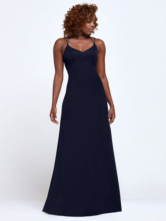 A woman with curly hair models Avery Austin’s navy 83045 A-Line floor-length dress with spaghetti straps, standing against a plain white background and gazing slightly to the side.