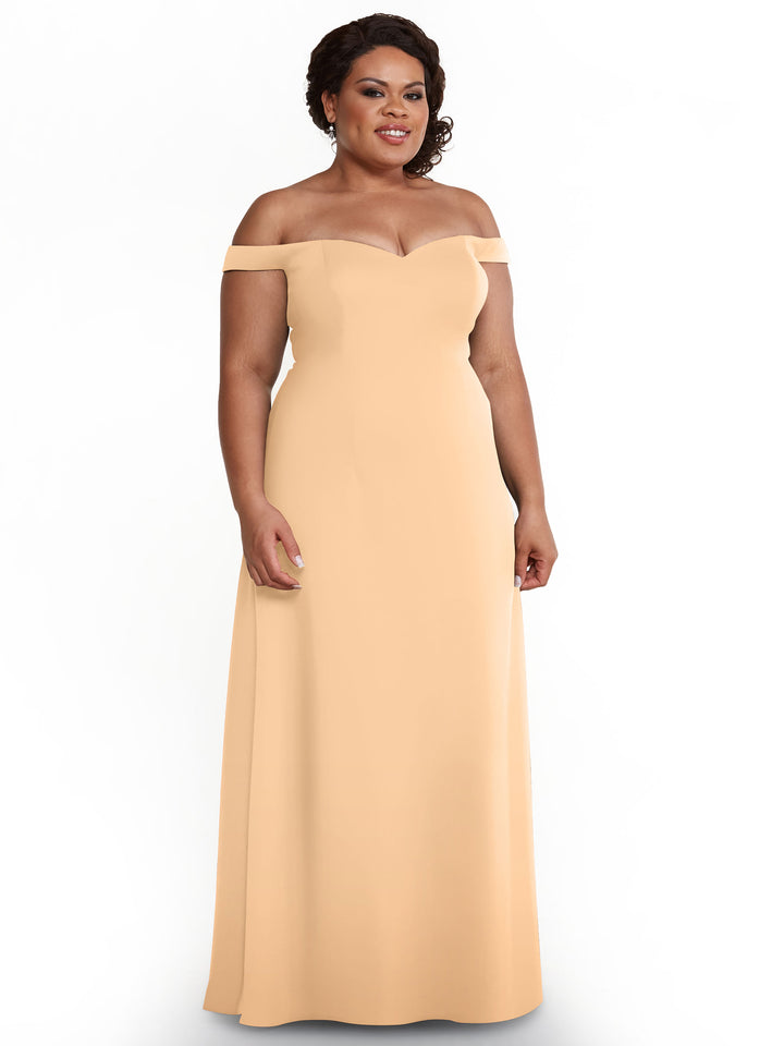 A woman models Avery Austin’s blush 83049 off-the-shoulder A-line dress, standing against a white background with a slight smile and her hands relaxed at her sides.