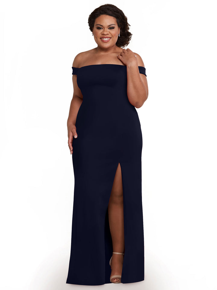 A woman smiles in a Navy Avery Austin 83052 off-the-shoulder bridesmaid dress with a thigh-high side slit, one hand touching her hair and the other at her side. She pairs the sheath dress with strappy heels against a white backdrop.