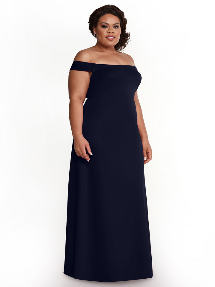 A woman models Avery Austin 83055, a navy floor-length off-the-shoulder gown with a subtle keyhole design, standing against a white background and smiling gently as she looks slightly to her left.