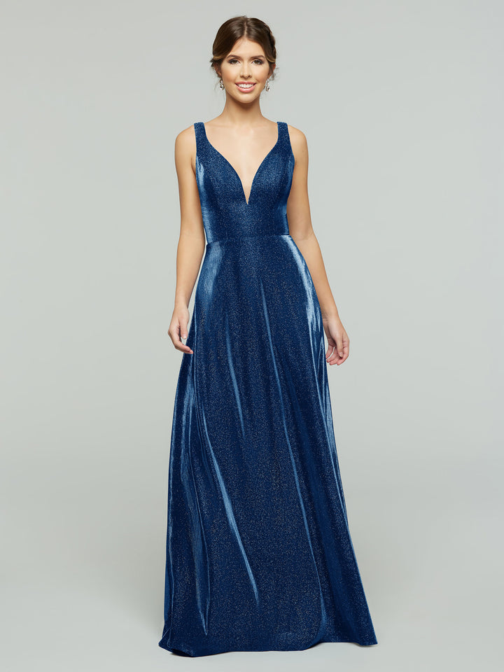 A woman stands smiling against a light gray background, wearing the averyaustin 84013 - Tried On Sample gown in Glitter Sapphire—a sleeveless, deep V-neck, floor-length dress with illusion cut-out sides and an elegant A-Line silhouette.