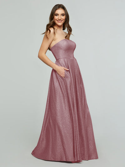A woman models the averyaustin 84014 - Tried On Sample, a sparkling, strapless mauve gown with a corseted back and pockets, smiling in the floor-length dress against a plain light gray background.