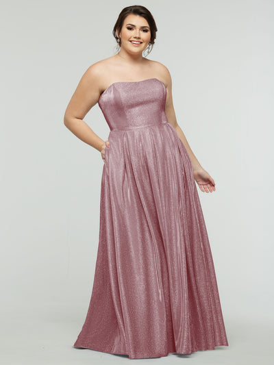 A woman smiles against a plain background, wearing the floor-length averyaustin 84014 - Tried On Sample gown in Glitter Mauve, featuring a strapless design, corseted back, and subtle shimmer that highlights her elegance.