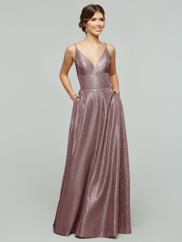 A woman models the averyaustin 84015 Tried On Sample, a sleeveless Glitter Rose Gold A-Line gown with deep V-neck and full-length skirt, standing with hands in pockets against a plain gray background.
