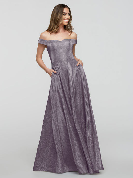 A woman models the averyaustin 84020 - Tried On Sample: an off-the-shoulder, floor-length, shimmery lavender A-Line gown with pockets, posing and smiling against a plain light gray background.